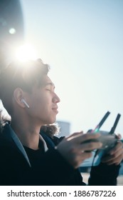 Young Asian Man Holding  Radio Remote Controller On The Street. Boy Flying A Drone Outside At Daytime. Quadcopter Operator Standing In Sunlight. Shallow Depth Of Field.