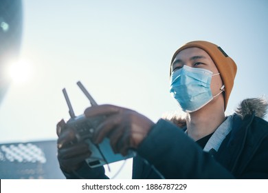 Young Asian Man Holding  Radio Remote Controller On The Street. Boy Flying A Drone Outside At Daytime. Quadcopter Operator Standing In Sunlight With Face Mask. Shallow Depth Of Field.