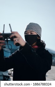 Young Asian Man Holding  Radio Remote Controller On The Street. Boy Flying A Drone Outside At Daytime. Quadcopter Operator Standing In Sunlight With Face Mask. Shallow Depth Of Field.