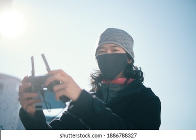 Young Asian Man Holding  Radio Remote Controller On The Street. Boy Flying A Drone Outside At Daytime. Quadcopter Operator Standing In Sunlight With Face Mask. Shallow Depth Of Field.