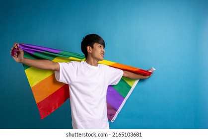 A Young Asian Man Holding A Rainbow Flag Against The Blue Color Background. Man With A Gay Pride Flag In Studio, With A Blank Space To The Right