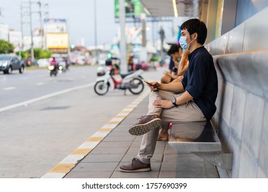 Young Asian Man Holding Smartphone And Waiting For Bus At Bus Stop In City Street And Wearing Face Mask Protective For Spreading Of Coronavirus(covid-19) Pandemic, New Normal Concept