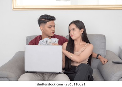A young asian man with a laptop on his lap is not amused by his materialistic girlfriend getting some of the money from their online business. A young wife holding multiple dollar bills. - Shutterstock ID 2365110021