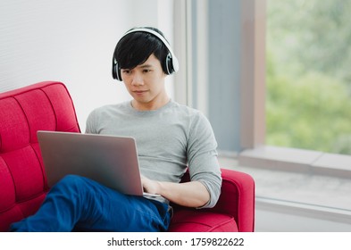 Young Asian Man With A Little Smile On Face Laying Down On Red Couch Using Laptop And White Headphone For Working Or Entertainment In Living Room