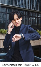 Young Asian Man, Looking At His Watch And Talking On The Phone Outside The Office. High Quality Photo