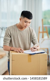 Young Asian Man Looking Information On Mobile Phone And Filling The Form On Cardboard Box While Standing At Home