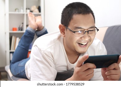 Young Asian man lying on cozy sofa, playing games on his smart phone, happy leisure activity