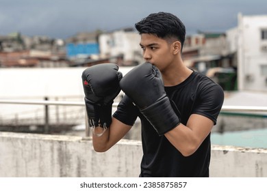 A young asian man makes a guard stance while practicing his boxing skills on the rooftop of a building. Shadowboxing training, wearing gloves, gritty urban scene. - Powered by Shutterstock