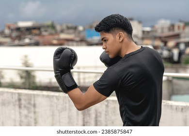 A young asian man makes an uppercut while practicing his boxing skills on the rooftop of a building. Shadowboxing training, wearing gloves, gritty urban scene.