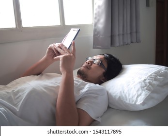 Young Asian Man With Mobile Smart Phone Lying Down On The Bed.