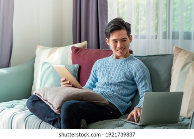 Young Asian Man On A Leisurely Day Sitting Home On Sofa Using Laptop And Tablet On The Couch.