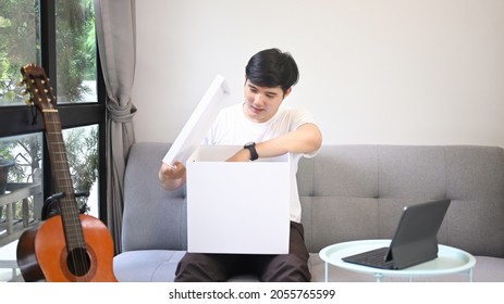 Young Asian Man Opening Gift Box While Sitting On Sofa.