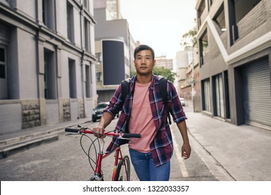 Young Asian man in a plaid shirt walking with his bicycle down an empty street in the city during his commute