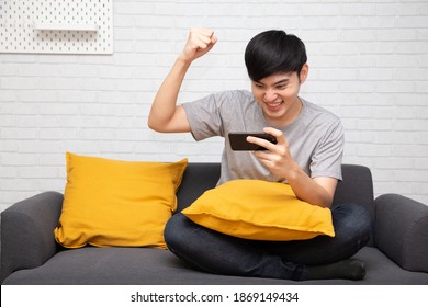 Young Asian Man Play Game On Mobile Phone Application And Raising His Arm Up With Celebrating Success On Sofa At Home