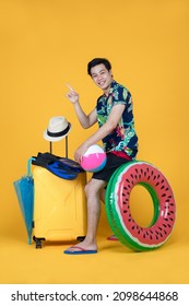 Young Asian man posing funny acting as happy tourist preparing for sea trip of summer vacation with outdoor sports equipment of beach ball, ring float, umbrella, hat, fins, and traveling luggage.
