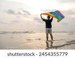 Young asian man with pride movement LGBT holding rainbow flag raise up for freedom. Demonstrate rights LGBTQ celebration pride Month Gay Pride Symbol. Standing on the sand sea beach during sunset