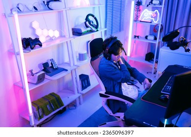 A young Asian man professional gamer wearing a jeans jacket sits on a chair and rubs a sore shoulder that suffering from stress playing a game or competition. Concept of pain from gaming and esports. - Shutterstock ID 2365096397