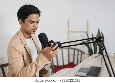 Young asian man radio host working, setting microphone, preparing to speak before recording podcast and live on social media. Technology of on-air online in broadcasting at home studio. - Powered by Shutterstock