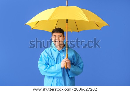 Young Asian man in raincoat and with umbrella on color background