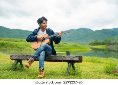 Young Asian man relax and enjoy outdoor lifestyle travel nature camping in forest mountain on summer holiday vacation. Handsome guy drinking coffee and playing guitar by the lake in the morning.