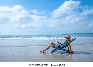 Young Asian man resting on sunbed on tropical beach. Happy guy sit on beach chair by the sea using smartphone for selfie or video call. Handsome male enjoy beach outdoor lifestyle on summer vacation