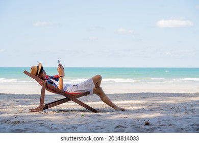 Young Asian man resting on sunbed on tropical beach. Happy guy sit on beach chair by the sea using smartphone for selfie or video call. Handsome male enjoy beach outdoor lifestyle on summer vacation - Powered by Shutterstock