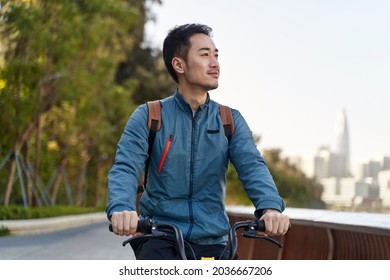 Young Asian Man Riding Bike On Riverfront Path In Public Park In Modern City