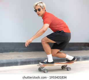 Young Asian Man Riding Skateboard On A City Street. Portrait Of Male In T-shirt And Black Short Pants Exercise Surf Skate Near Sidewalk In Urban. Trendy Outdoor Extreme Sport Recreation In Asia. 