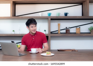 A young Asian man sat and worked from home in the dining room. Which is decorated in wood and looks modern, he is using a smartphone and laptop while drinking coffee and eating sandwiches. - Powered by Shutterstock