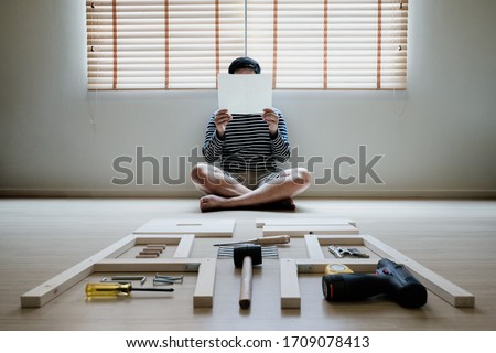 Young asian man self assembly wooden furniture and read blank white paper to put instruction