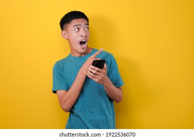 Young Asian Man With Shock Face Holding A Mobile Phone, Pointing And Looking At Copy Space, Isolated On Yellow Background