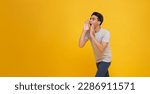 Young Asian man shouting open mouths raising hands screaming announcement to empty space aside isolated on yellow studio background.
