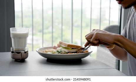 A Young Asian Man Sits At The Dining Room Table Full Of Food, He Dunks The Food On The Plate And Eats It, Relishing It.