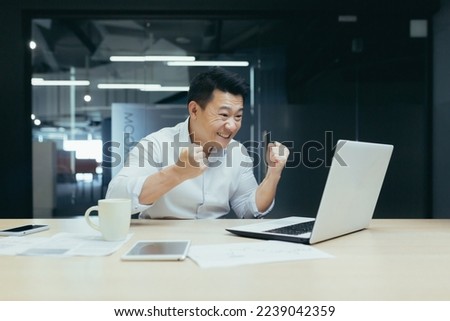 A young Asian man sits in the office at the table, during working hours he watches a football match, sports competitions,makes bets, cheers and rejoices on his laptop.