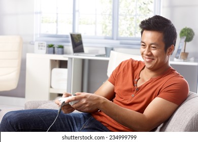 Young Asian Man Sitting In Armchair At Home, Playing Video Game, Smiing.