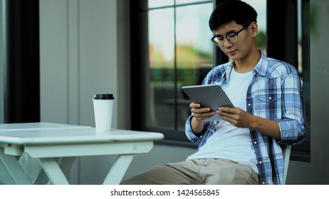 Young asian man sitting on chair, he use tablet in hand and a drinking beverage placed on table, copy space for insert text - Powered by Shutterstock