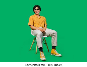 Young Asian Man Sitting On Chair With Laugh And Smile Expression. Wearing Black Sun Glasses. Isolated On Green Background With Clipping Path