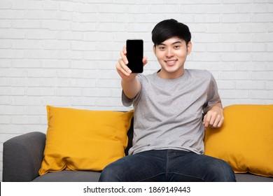 Young Asian Man Sitting On Sofa And Showing Present Mobile Phone Application At Home