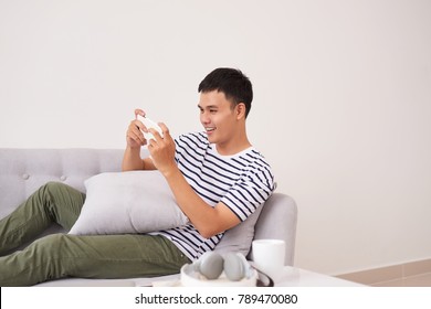 Young Asian Man Sitting On Sofa And Playing Games On Phone At Home