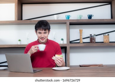 A young Asian man sitting and working from home in the dining room. Which is decorated in wood and looks modern, he is using a laptop while drinking coffee and eating sandwiches. - Powered by Shutterstock