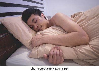 Young Asian man smiling and hugging the pillow while sleeping in his bed at home.