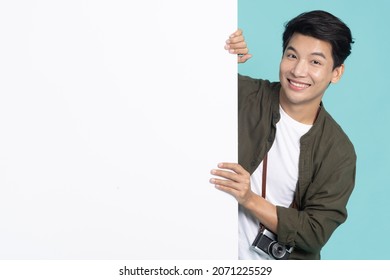 Young Asian Man Is Standing Behind The White Blank Banner Or Empty Copy Space Advertisement Board On Green Background, Looking At Camera