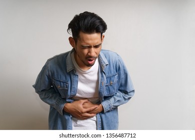 Young Asian Man Standing And Holding His Tummy Where He Feels Pain Due To Stomachache Problem. His Expression On Face Show How Much He Hurt. Seen In White Background. Sickness Portrait Concept.