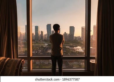 Young asian man standing and looking through window in a hotel. Sunrise over modern buildings in Songdo central park at Incheon, South Korea - Powered by Shutterstock