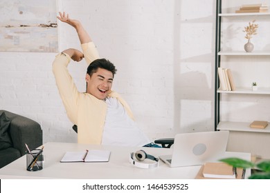 Young Asian Man Stretching And Smiling While Sitting At Desk Near Laptop