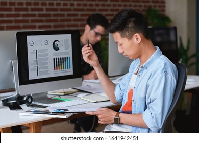 Young asian man student working in office reading financial document with graph information studying finance sitting by desk with computer