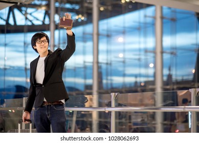 Young Asian Man With Suitcase Luggage Raising Hand For Greeting With His Friend In The International Airport Terminal, Business Travel Concept