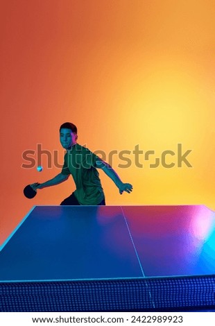 Young Asian man, table tennis athlete preparing for return, hit ball in neon light against warm yellow gradient background. Dynamic gel portrait. Concept of professional sport, healthy lifestyle. Ad