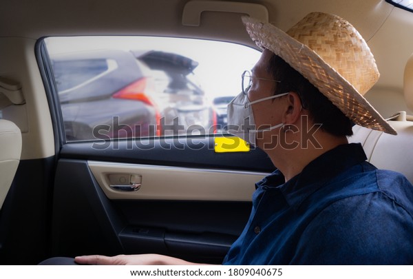 Young Asian man takes a taxi and looks out\
traffic jams wearing a sterile medical mask. A man sits on the back\
seat of the taxi and takes a ride during coronavirus pandemic.\
Social distance concept.
