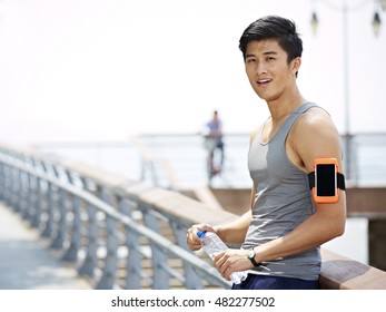 Young Asian Man Taking A Break With A Bottle Of Water In Hands During Outdoor Exercise